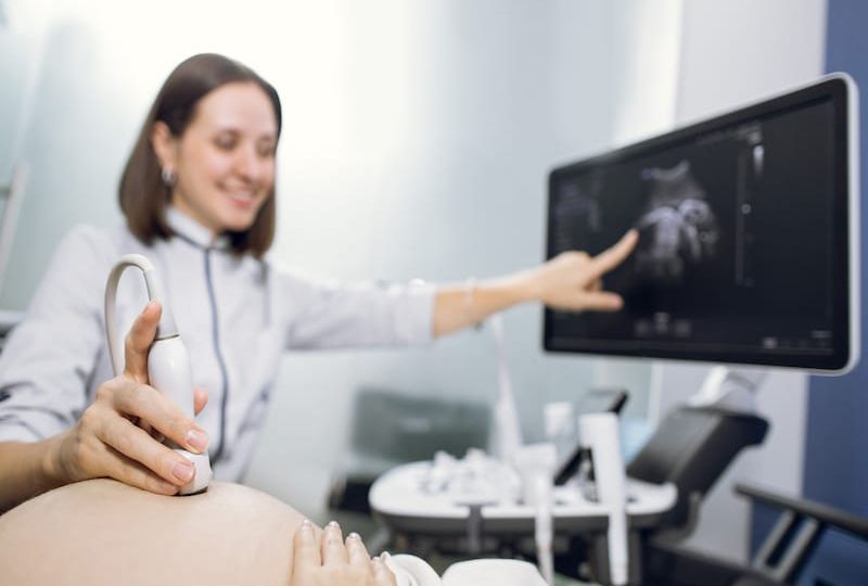 Cropped shot of a pregnant woman during ultrasound scanning at the fertility clinic. Female doctor pointing at the screen of ultrasound machine. Focus on ultrasound transducer on the pregnant belly