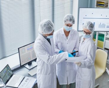 Group of doctors in white coats masks and gloves discussing medical document together while standing at office