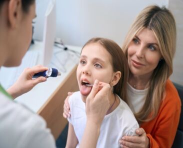 Female ear, nose and throat specialist examining throat of little girl sitting on mothers knees with complains of sore throat, health check-up