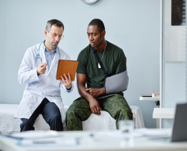 Young Black soldier with broken arm wearing sling sitting on medical couch having doctors appointment