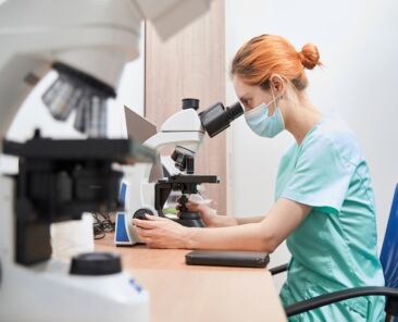 Close-up photo of female scientist in uniform sitting at the table, working in the laboratory, using microscope